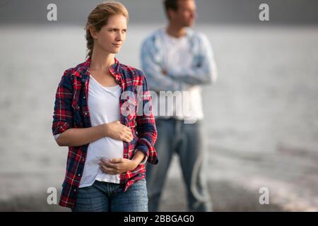 Porträt einer schwangeren Frau am Strand, die sanft ihren Bauch hält und ihren Mann im Hintergrund steht Stockfoto