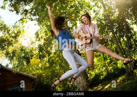 Zwei glückliche Frauen in der Mitte des Erwachsenen springen in der Luft mit einer Gitarre Stockfoto