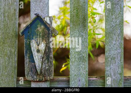 Handgefertigte, hausförmige Holzplatte mit Herzform an einem Gartenzaun. Baum mit leuchtend grünen Blättern im sonnenbeleuchteten Hintergrund. Haus, Gartenkonzept Stockfoto