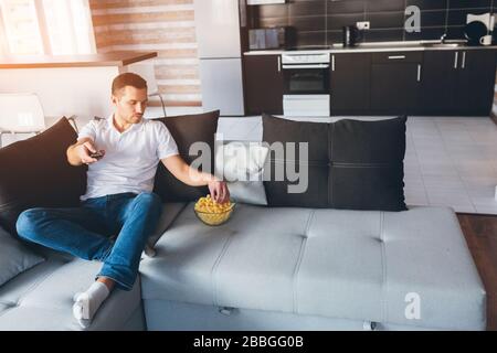 Der junge Mann gucken in seiner eigenen Wohnung fernsehen. Der konzentrierte Kerl hält die Fernbedienung in der Hand und bekommt einen Snack aus der Schüssel. Sehen Sie sich Filme oder fernsehsendungen allein im Zimmer an Stockfoto
