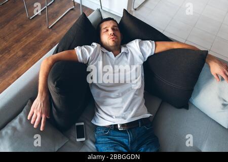 Der junge Mann gucken in seiner eigenen Wohnung fernsehen. Allein auf der Couch müde und erschöpft schlafen. Tagträumen. Gewöhnlicher Kerl ruht sich aus und entspannt sich. Hände liegen auf Schwarz Stockfoto