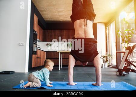 Vater führt einen Handstand durch, während sein Baby auf dem Boden krabbelt Stockfoto
