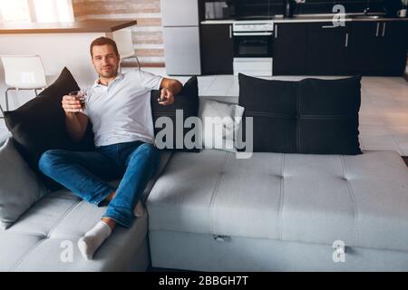 Der junge Mann gucken in seiner eigenen Wohnung fernsehen. Guy sitzt auf dem Sofa und schaut mit einem Glas Alkohol in der Hand auf den fernseher. Fröhlicher positiver junger Mann trinkt Whisky oder Stockfoto