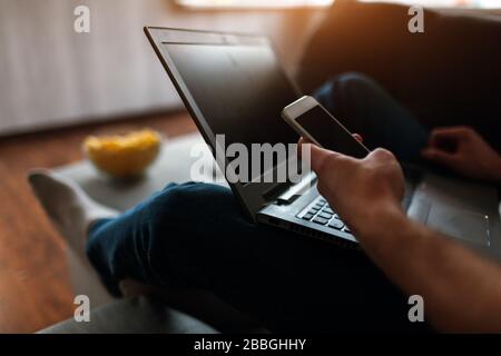 Der junge Mann gucken in seiner eigenen Wohnung fernsehen. Schneiden Sie die Ansicht und Nahaufnahme des Mannes, der das Smartphone in der Hand hält. Laptop an seinen Beinen. Film oder Programm ansehen Stockfoto