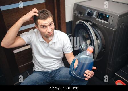 Der konzentrierte Kerl in der Badewanne hält flüssiges Pulver und schaut auf die Kamera. Kratzen Sie den Kopf und denken Sie an eine Flasche in der Hand. Richtig verwenden und Stockfoto