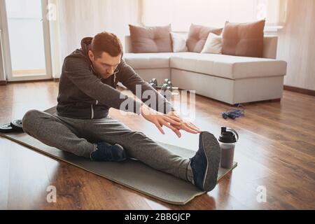 Der junge normale Mann geht zu Hause für Sport ein. Neuling oder Anfänger, die sich mit den Händen bis zu einem Bein Strecken. Alleine in leerer Wohnung trainieren. Versuchen Sie es Stockfoto