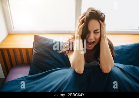 Young beautiful woman in morning bed at home. Sit and hold hands on head. Scream because of pain and stress. Depression and annoying condition. Stockfoto