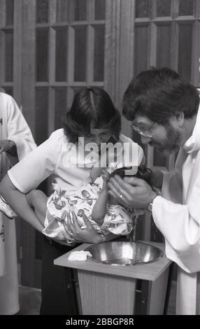 1970er Jahre, historisch, in einer Kirche, eine Mutter, die ihr kleines Kind hält, während es von einem gekleideten Priester getauft wird, South London, England, Großbritannien. Die Taufe ist ein christlicher Einlassritus, ein Sakrament, eine Verordnung Jesu Chris Stockfoto