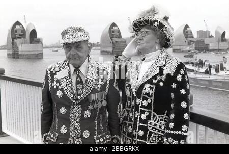 1984 tragen Londons Perlkönig und Königin ihre traditionellen Outfits. Jacken mit Perlmuttknöpfen, die zusammen an den Royal Docks stehen, bei der Eröffnung der neuen Thames Barrier, South London, England, Großbritannien durch Königin Elizabeth II..stellvertretend für Londons Arbeiterkultur, "Pearlies", wie sie auch bekannt sind, Entstanden aus den Coster Kings und Queens, die als Führer der Londoner Straßenhändler gewählt wurden. Stockfoto