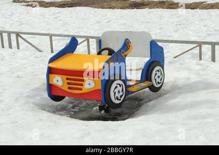 Kinderschaukel in Form eines Autos auf dem Spielplatz vor dem Hintergrund winterlicher Schneeverwehungen. Konzept für Kinderunterhaltung und Straßenspiele. Stock-Foto für Web und Druck mit leerem Platz für Text. Stockfoto