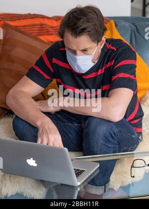 MID 40er Männer arbeiten von zu Hause aus mit Apple-Laptop und tragen Gesichtsmaske. Stockfoto