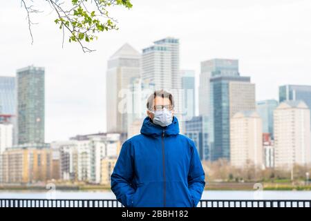 Männer aus Mitte 40 tragen eine blaue Jacke und eine Gesichtsmaske, die auf der Themse entlang spazieren, mit Londons Finanzbezirk und Canary Wharf im Hintergrund. Stockfoto