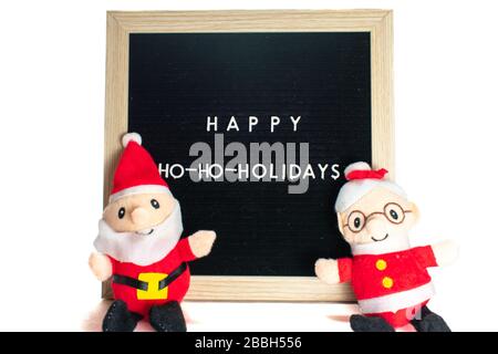 Ein schwarzes Schild mit weißen Buchstaben in einem Birch-Rahmen, das frohe Feiertage mit dem Weihnachtsmann sagt, und Frau Claus vor ihm Stockfoto