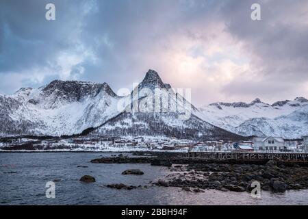 Skandinavisches Dorf an der Küste mit schneebedeckten Bergen im Hintergrund bei Mefjord Brygge, Senja Island, Norwegen Stockfoto