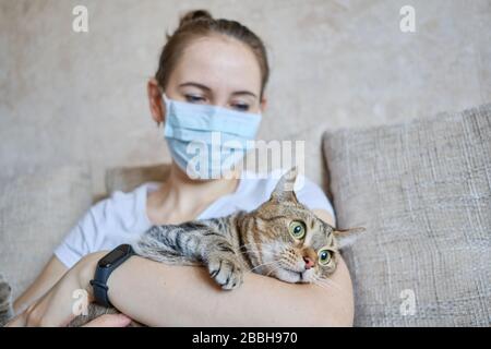 Das Mädchen in einer medizinischen Maske befindet sich zu Hause in Selbstisolation und kümmert sich um die Katze. Nahaufnahme. Stockfoto