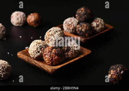 Gesunde Energiekugeln aus Nüssen, Haferflocken und Trockenfrüchten mit Kokosnuss, Flachs und Sesamsamen auf Holz-Kokosnuss-Platten auf schwarzem Grund, horizontal Stockfoto