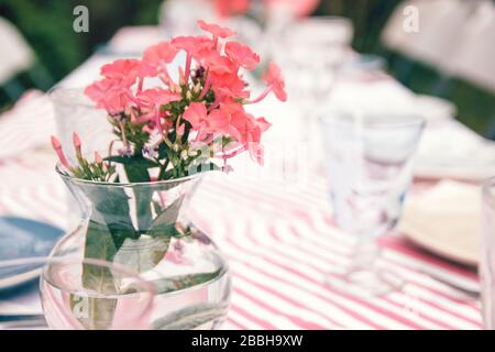 Ländlicher Hochzeitstisch mit Korallenblumen in einer Glasvase auf einem gestreiften Tischtuch. Spezielle Ereignistabelle eingerichtet. Frische Blumendekoration. Stockfoto