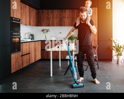 Der junge Vater saugt den Boden der Wohnung mit seinem Baby auf dem Hals Stockfoto