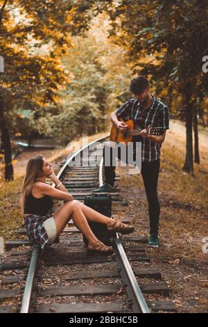 Ein junges Liebespaar verpasste den Zug. Song mit Gitarre auf den Schienen im Herbstwald, der auf den nächsten Zug wartet. Stockfoto