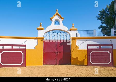 Große rote Tore in der Stierkampfarena, weiße Wände mit orangefarbener Dekoration, zwei rote Schranken und sandiger Boden. Isla Mayor, Spanien Stockfoto