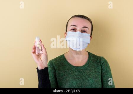 Weiße kaukasische Erwachsene Frau im Pullover und mit medizinischer Maske auf dem Gesicht hält einen Krug mit antiseptischem Gel in der Hand. Auf beigefarbenem Hintergrund. Kopierbereich. Stockfoto