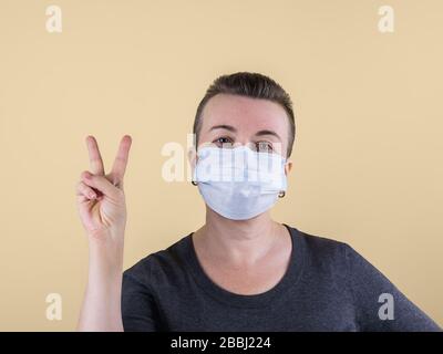 Sehr kurzhaarige kaukasische Erwachsene Frau im Pullover und mit medizinischer Maske auf dem Gesicht zeigt Siegeshandgeste. Auf beigefarbenem Hintergrund. Kopierbereich. Stockfoto