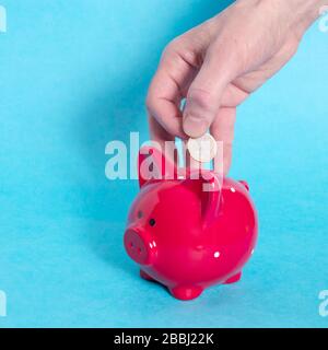 Männliche Hand wirft einen Euro in eine rote, piggy Bank mit einem weißen Punkt auf blauem Hintergrund. Cocept. Kopierbereich. Stockfoto