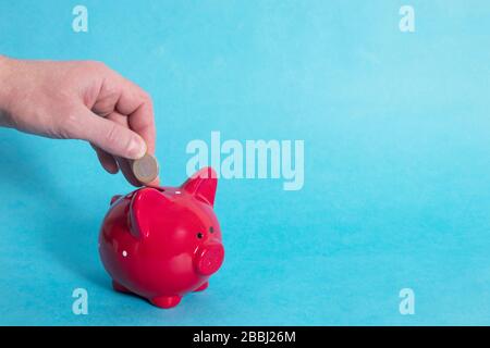 Männliche Hand wirft einen Euro in eine rote, piggy Bank mit einem weißen Punkt auf blauem Hintergrund. Cocept. Kopierbereich. Stockfoto