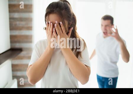 Junge Frau und Mann während der Quarantäne im Zimmer. Frau steht vor und schreit. Ihr Gesicht mit den Händen bedecken. Mann schreit und schreit Frau an. Emotional Stockfoto