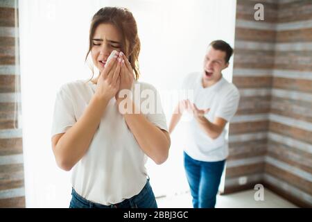 Junge Frau und Mann während der Quarantäne im Zimmer. Guy schreit seine Frau an. Sie weinte und hielt weißes Gewebe auf Wange. Abusing und Eigenheimagrevität. Stockfoto
