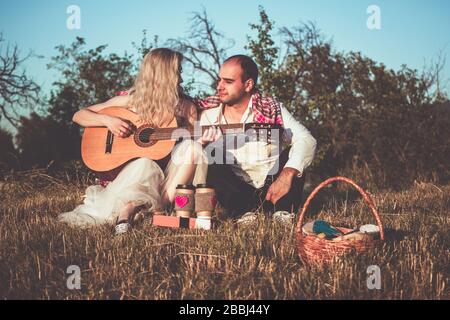 Ein romantisches Paar umarmt, während es bei Sonnenuntergang Picknick machen kann. Mann in weißem Hemd und attraktive junge Frau, die Gitarre spielt, sitzen zusammen Stockfoto