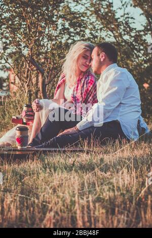 Ein romantisches Paar umarmt, während es bei Sonnenuntergang Picknick machen kann. Mann in weißem Hemd und attraktive junge Frau sitzen zusammen auf Gras auf dem Stockfoto