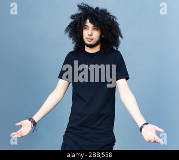 Ein junger, modisch gekleideter Mann mit einer Afro Frisur und einem Bart in einem coolen Outfit, der an einer grauen Wand posiert. Er breitet seine Arme überraschend aus Stockfoto