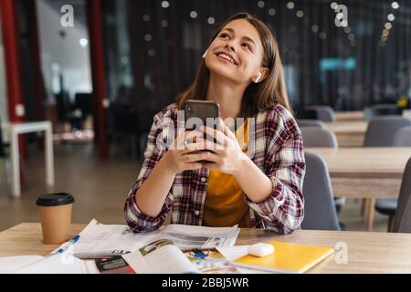 Bild eines glücklichen netten Mädchens mit drahtlosem Ohrhörer und Handy während der Hausaufgaben in der College-Bibliothek Stockfoto