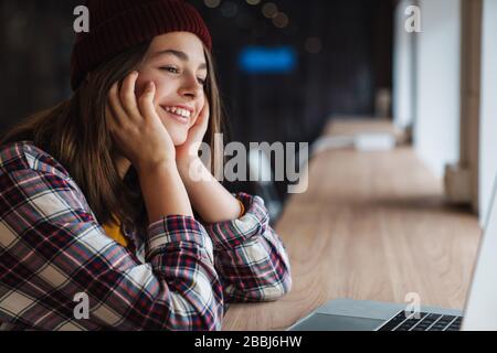 Bild des fröhlichen kaukasischen Mädchens in einem Hut lächelnd und mit Laptop beim Sitzen in der College-Bibliothek Stockfoto