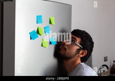 Junger hispanischer Mann mit Bart und Brille, überwältigt von Erinnerungen, die auf dem Kühlschrank aufgeklebt wurden Stockfoto