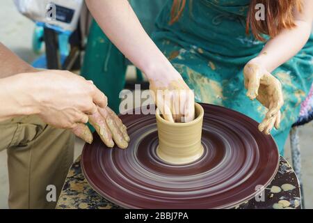 Die Hände eines Meisters und eines Studenten auf einem Potters Rad sculpten einen Pitcher aus Ton. Selektive Fokussierung auf die Hände Stockfoto