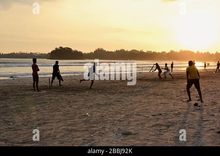 Junge Jungen spielen Cricket-Spiel an einem Strand. Silhouetten von jungen Jungen, die am Strand von Sri Lanka Spaß haben. Goldenes Licht bei Sonnenuntergang mit Sonnenstrahlen in Weligama Stockfoto