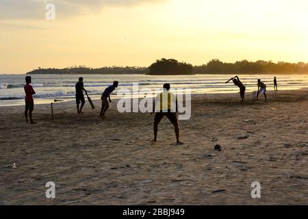 Junge Jungen spielen Cricket-Spiel an einem Strand. Silhouetten von jungen Jungen, die am Strand von Sri Lanka Spaß haben. Goldenes Licht bei Sonnenuntergang mit Sonnenstrahlen in Weligama Stockfoto