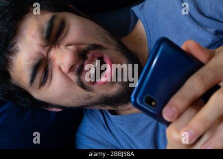 Bärtiger hispanischer junger Mann in einem Wohnzimmer mit seinem Smartphone und zeigt negative Reaktionen wie Wut, Frustration, Langeweile, Wut und anderes c Stockfoto