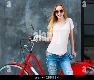 Streetstyle. Wunderschönes blondes Mädchen im trendigen, weißen Outfit, das mit einem trendigen roten Fahrrad im Freien posiert. Stockfoto