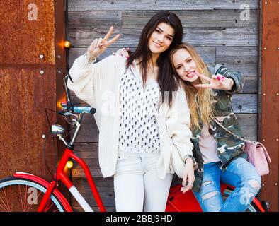 Zwei trendige Mädchen mit rotem Vintage-Rad auf altem Holzhintergrund zeigen ein Zeichen des Sieges. Foto Mit Tonnagentoning. Modernes Jugend-Lifestyle-Konzept. Stockfoto