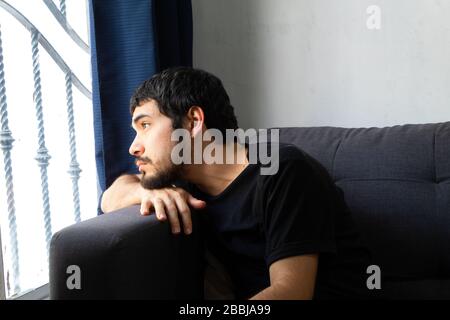 Bärtiger hispanischer junger Mann, der auf einem Sofa sitzt und sehr nachdenklich auf ein Fenster blickt Stockfoto