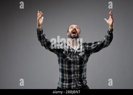 Mann im kariertem Hemd mit ausgestreckten Armen, Blick auf den Himmel, als ob er bete oder Erfolge feierte. Halblanges Vorderporträt gegen Grau B. Stockfoto