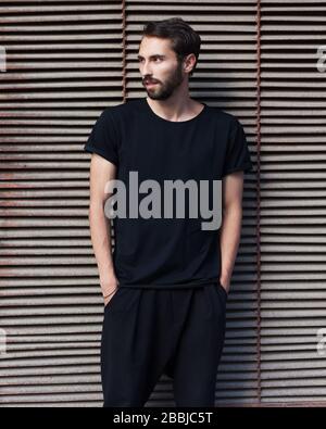 Ein bärtiger Hüftkämpfer in einem schwarzen T-Shirt und einer Hose, die am Nachmittag gegen eine rostige Metallwand posiert. Stockfoto