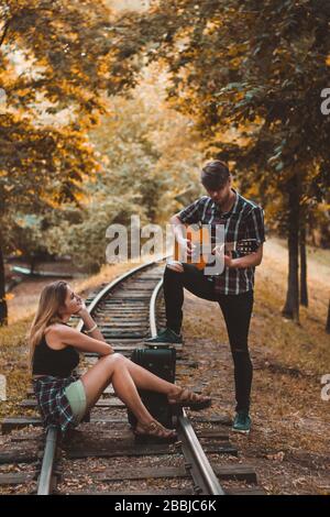 Ein junges Liebespaar verpasste den Zug. Song mit Gitarre auf den Schienen im Herbstwald, der auf den nächsten Zug wartet. Stockfoto