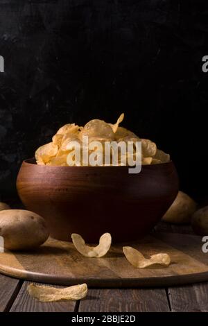 Kartoffelchips in einer Schüssel auf rustikalem Holzhintergrund mit ganzen Kartoffeln. Salzige Knäckchen auf einem Tisch. Stockfoto