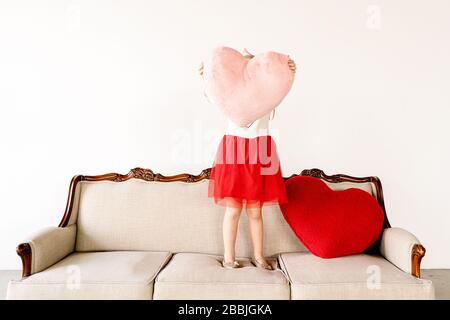 Kleines Mädchen mit großen rosa Herz Kissen auf Couch drinnen Stockfoto