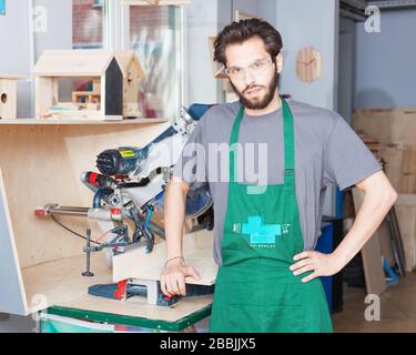 Porträt eines bärtigen Handwerkers mit Elektrowerkzeug in großzügiger Werkstatt und Blick auf die Kamera Stockfoto