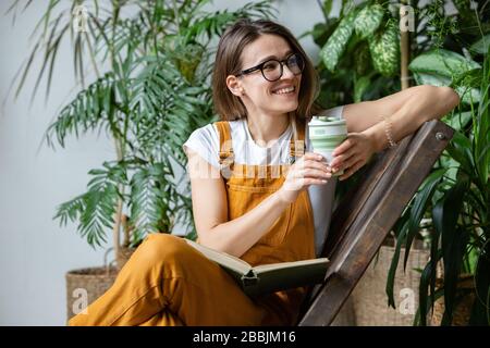 Weibliche europäische Gärtnerin, die Overalls trägt, nach der Arbeit ruht, auf Holzstuhl im heimischen Gewächshaus sitzt, wiederverwendbaren Kaffee-/Teebecher beiseite hält Stockfoto